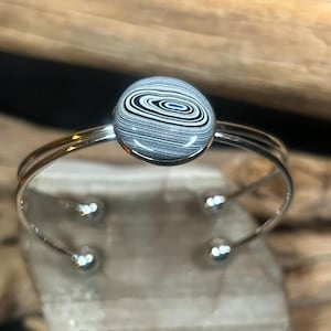 May include: A silver-toned cuff bracelet featuring a round, marbled stone with a black, white, and blue swirl pattern. The bracelet has a double-band design and small, decorative silver beads at the ends.