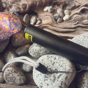 May include: A cylindrical black laser pointer with a yellow warning label and a light gray woven wrist lanyard. The device is positioned among various smooth gray river stones and a single iridescent rock with orange and purple flecks.