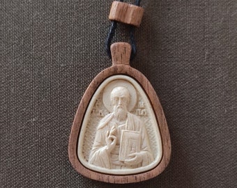 May include: A wooden pendant with a carved image of a saint holding a book. The pendant is on a brown cord.