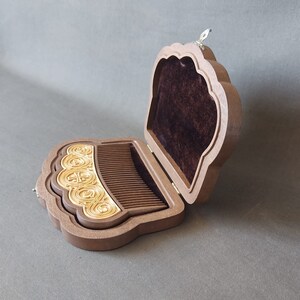 Puede incluir: Un peine de madera con un dise&ntilde;o dorado est&aacute; en una caja de madera abierta con un forro de terciopelo marr&oacute;n. La caja tiene una tapa con bisagras con un dise&ntilde;o decorativo.