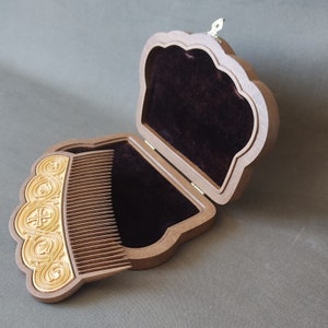 Puede incluir: Un peine de madera con un dise&ntilde;o dorado est&aacute; en una caja de madera abierta con un forro de terciopelo marr&oacute;n oscuro. La caja tiene una tapa con bisagras y un peque&ntilde;o pestillo de metal.