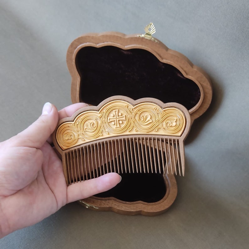 Puede incluir: Un peine de madera con un dise&ntilde;o decorativo y ornamentado en una caja de madera. El peine tiene un acabado dorado y presenta una cruz y motivos florales.