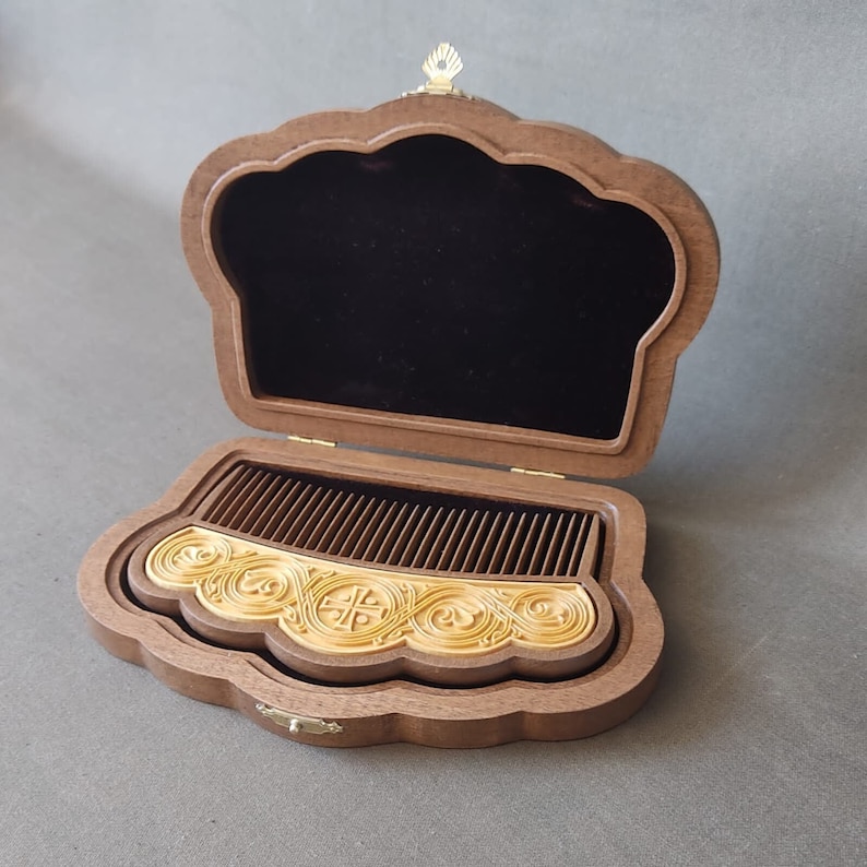 Puede incluir: Una caja de madera con una tapa con bisagras, la tapa est&aacute; abierta y revela un peine dorado con intrincados tallados. La caja tiene forma de concha de vieira y tiene un peque&ntilde;o pomo dorado en la parte superior.