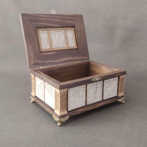 May include: A wooden box with a hinged lid and decorative carvings. The box is open and the inside is lined with a light-colored material. The box has four brass feet.