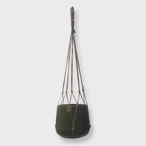 May include: A dark olive green ceramic plant pot suspended by a brown cord macrame hanger. The hanger features a series of knotted cords that converge at a top loop, supporting the rounded, smooth-surfaced pot from the base and sides.
