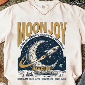 May include: White t-shirt featuring a graphic design with the words "MOON JOY" in gold lettering. The design includes a crescent moon, a rocket ship, and the text "CELEBRATING HUMANITY'S RETURN TO THE LUNAR FRONTIER" and "ARTEMIS II".