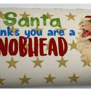 Op de afbeelding: Een verpakte chocoladereep met een feestelijk ontwerp. De tekst luidt "Santa Thinks you are a KNOBHEAD" in groen, blauw en rood. De kerstman is rechts afgebeeld. Gouden sterren zijn verspreid over de witte achtergrond.