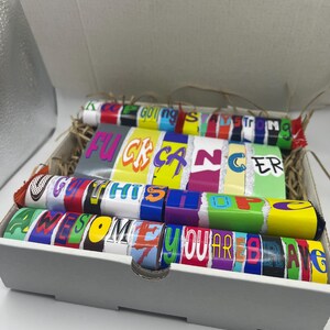 Puede incluir: Una caja de regalo gris llena de coloridas barras de caramelo. Los envoltorios muestran frases como "Keep Going Strong", "F*ck Cancer", "Get Thru This", "Awesome" y "You Are Brave". La caja est&aacute; forrada con paja.