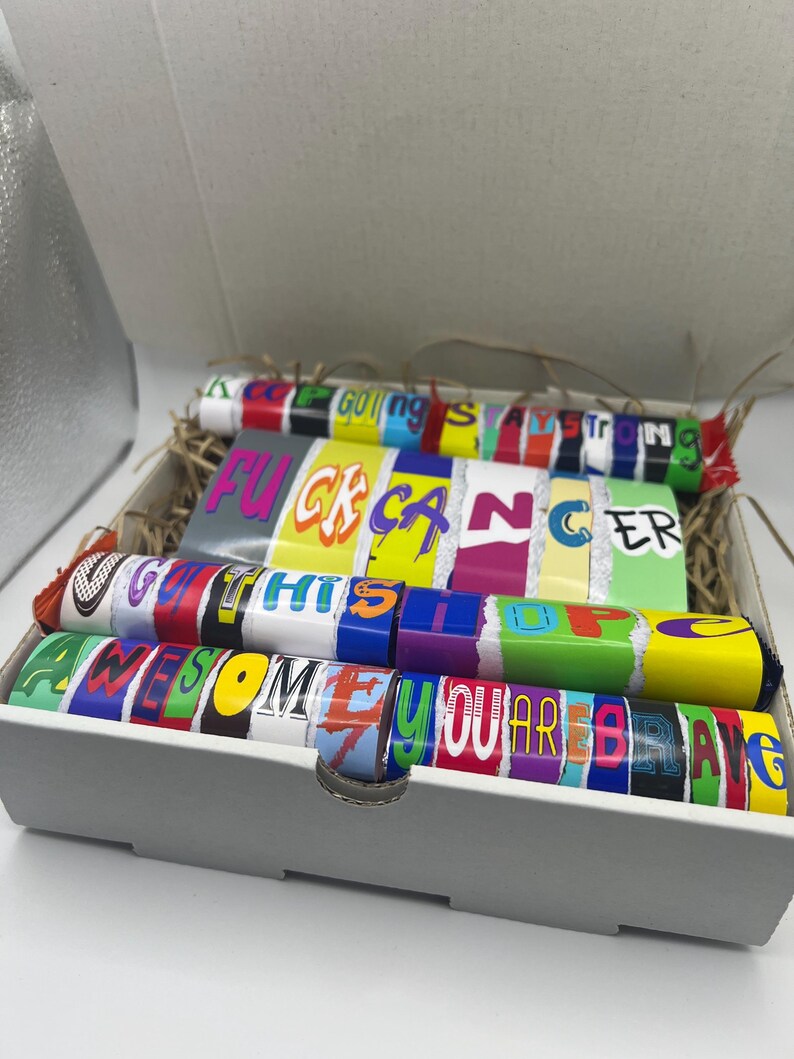 Puede incluir: Una caja de regalo gris llena de coloridas barras de caramelo. Los envoltorios muestran frases como "Keep Going Strong", "F*ck Cancer", "Get Thru This", "Awesome" y "You Are Brave". La caja est&aacute; forrada con paja.