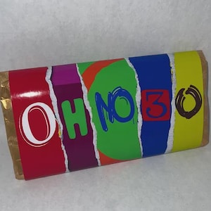 Op de afbeelding: Een rechthoekige verpakking van een chocoladereep met de tekst "OH NO 30" in een kleurrijk, gescheurd papierontwerp. De verpakking heeft rode, paarse, groene, blauwe en gele secties met gouden folieranden.
