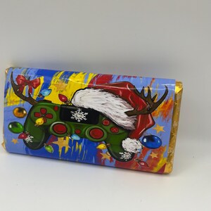 Puede incluir: Una barra de chocolate envuelta con un diseño navideño. El envoltorio presenta un mando de videojuegos verde con cuernos de reno, un gorro de Papá Noel y luces navideñas. El fondo es azul y amarillo con estrellas doradas.