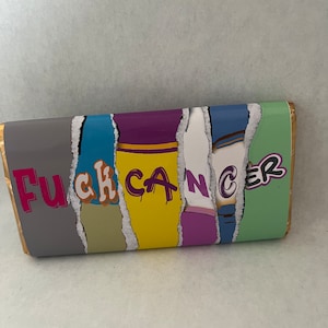 Puede incluir: Envoltorio de barra de chocolate con las palabras "Fuck Cancer" en un dise&ntilde;o colorido de papel rasgado. El envoltorio tiene un borde dorado y un fondo gris.