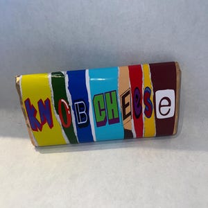 May include: A rectangular package with the text "KNOB CHEESE" in a colourful, layered design. The package features a yellow, green, blue, and red colour scheme, with a brown border.