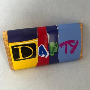 Op de afbeelding: Een rechthoekige verpakking van een chocoladereep met het woord "DAFTY" in vette letters. De verpakking is verdeeld in secties van geel, rood, donkerblauw, groen en lichtblauw. De letters zijn in verschillende kleuren en stijlen.