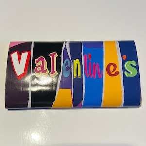 Op de afbeelding: Een kleurrijke Valentijnsdag-thema geschenkdoos met het woord "Valentine's" in verschillende lettertypen en kleuren. De doos heeft een gescheurd papierontwerp met een mix van paarse, zwarte, blauwe en gele secties.
