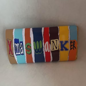 May include: A rectangular object with the text "Xmas Wanker" in colorful, block letters. The background is a series of vertical stripes in various colors, including blue, orange, and brown. The object appears to be a decorative item.