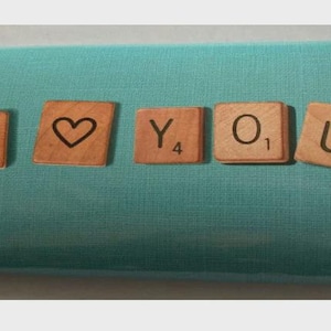Op de afbeelding: Een turquoise geschenkdoos met houten Scrabble-tegels met de tekst "I ❤️ YOU". De tegels zijn lichtbruin met zwarte letters en een hartsymbool. De doos is verpakt in turquoise papier.