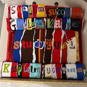May include: A box of assorted chocolate bars with colourful, torn paper wrappers. Each bar has a different message, including "Smartie", "Success", "Good Luck", "Believe", "Study Fuel", "Keep Calm", and "You Got This".