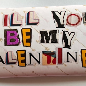 Op de afbeelding: Een gevouwen kaart met de woorden "WILL YOU BE MY VALENTINE?" in collage-stijl. De letters zijn uit verschillende materialen en kleuren geknipt, met een vraagteken aan het einde. De achtergrond heeft een patroon van pijlen.