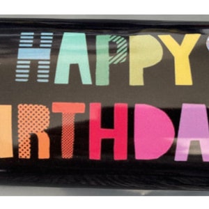 Op de afbeelding: Een zwarte chocolade reep met een gouden wikkel. De wikkel heeft een kleurrijke "Happy Birthday" boodschap met sterren en een ballon.