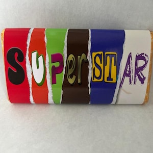Op de afbeelding: Een chocoladereep met het woord "SUPERSTAR" erop gedrukt in kleurrijke, gescheurde en overlappende letters. De letters zijn in rood, oranje, groen, bruin, blauw en paars.