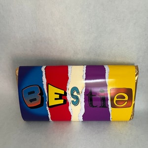 May include: A candy bar wrapped in colorful paper with the word "Bestie" in a playful font. The wrapper features blue, red, cream, purple, and yellow sections, creating a vibrant and eye-catching design.