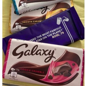 May include: Three Galaxy chocolate bars with different flavors: Cookie Crumble, Smooth Caramel, and Salted Caramel. The Cookie Crumble bar has a pink and white wrapper with a close-up of the chocolate and cookie pieces. The Smooth Caramel bar has a purple wrapper with the text "Missed the most famous star on the planet, Rose, 79". The Salted Caramel bar has a blue and white wrapper with a close-up of the chocolate and caramel pieces.