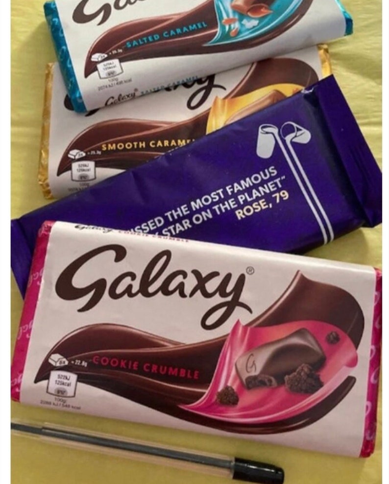 May include: Three Galaxy chocolate bars with different flavors: Cookie Crumble, Smooth Caramel, and Salted Caramel. The Cookie Crumble bar has a pink and white wrapper with a close-up of the chocolate and cookie pieces. The Smooth Caramel bar has a purple wrapper with the text "Missed the most famous star on the planet, Rose, 79". The Salted Caramel bar has a blue and white wrapper with a close-up of the chocolate and caramel pieces.