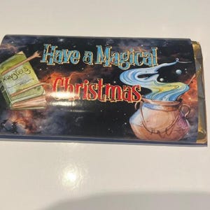 Op de afbeelding: Een ingepakt cadeau met een donkere achtergrond. Het heeft een ketel met kleurrijke rook, een boek met het label "Monsters" en de tekst "Have a Magical Christmas". Het cadeau heeft gouden folieranden.