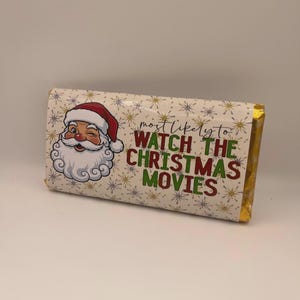 Op de afbeelding: Een verpakte chocoladereep met een knipogende kerstman-afbeelding. De tekst "most likely to watch the Christmas movies" is in groen en rood gedrukt op een crèmekleurige achtergrond met gouden folieranden.