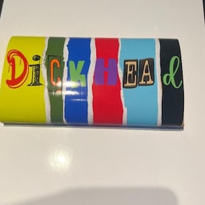 Op de afbeelding: Een rechthoekig object met het woord "Dickhead" in kleurrijke, gevarieerde lettertypen. De achtergrond is verdeeld in verticale strepen van geel, groen, blauw, rood, lichtblauw en zwart, met een gescheurd papiereffect.