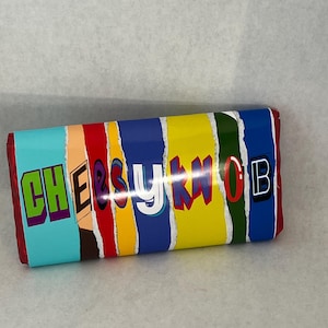 May include: A rectangular candy bar wrapper with the text "CHEESYKNOB" in colourful block letters. The wrapper has a design of vertical stripes in various colours, including blue, red, yellow, and green.
