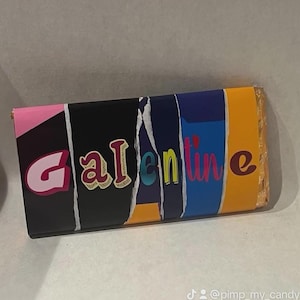 Op de afbeelding: Een wikkel van een chocoladereep met het woord "Galentine" in kleurrijke, gestileerde tekst. De wikkel heeft een gescheurd papierontwerp met roze, zwarte, blauwe en oranje secties. De chocoladereep is verpakt in goudfolie.