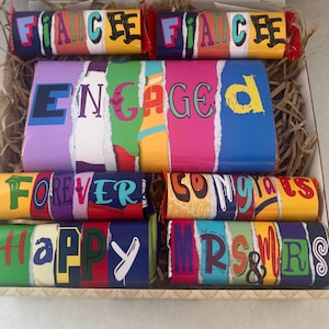 May include: A gift box containing candy bars with celebratory messages. The bars display the words "FIANCEE", "ENGAGED", "FOREVER", "CONGRATS", "HAPPY", and "MR & MRS" in vibrant colours and fonts. The box is lined with shredded brown paper.