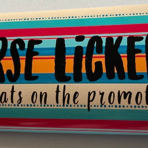 Pode incluir: Uma caixa de presente retangular com um padrão listrado colorido em vermelho, azul, laranja e verde-azulado. O texto preto em negrito diz "ARSE LICKER" acima de "Congrats on the promotion" em uma fonte menor.