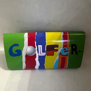 May include: A rectangular green chocolate bar wrapper displaying the word GOLFER in colourful, stylised lettering. The letter O is substituted with a white golf ball graphic. The design incorporates vertical, torn-paper style stripes in red, blue, and yellow.