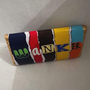 May include: A candy bar with a colorful wrapper featuring the word "WANKER" in a playful font. The wrapper has a navy blue section, followed by red, brown, yellow, light blue, and orange sections. The candy bar is wrapped in gold foil.