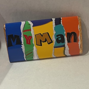 Op de afbeelding: Een rechthoekige chocoladereepverpakking met de woorden "MY Man" in zwarte en gouden letters. De verpakking is verdeeld in kleurrijke secties van blauw, groen, geel, turquoise en oranje, met een gescheurd papiereffect.