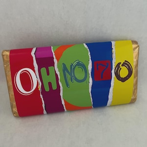 Op de afbeelding: Een kleurrijke wikkel van een chocoladereep met de woorden "OH NO 70". De wikkel heeft een ontwerp met rode, paarse, groene, blauwe en gele secties. De chocoladereep is verpakt in goudfolie.