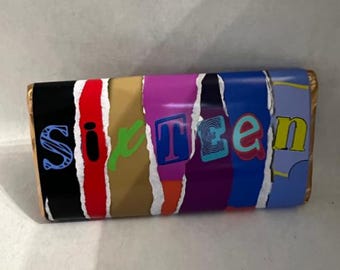 May include: A rectangular candy bar wrapper featuring a collage of vertical, torn-edge strips in black, red, gold, purple, dark blue, and light blue. The word SIXTEEN is printed across the center in varied, colorful, and stylized fonts.