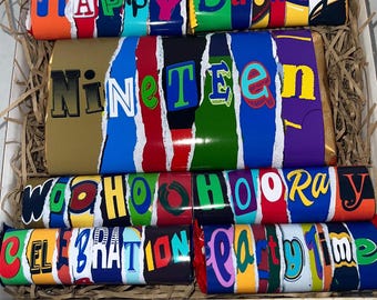 May include: A gift box filled with chocolate bars. The bars are wrapped in colorful paper with phrases like "HAPPY Birthday", "Nineteen", "Woohoo Hooray", "Celebration", and "Party Time". The text is in various colors and fonts.