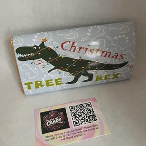 Op de afbeelding: Een snoepreepverpakking met een lichtblauwe achtergrond met een met een kerstboom versierde dinosaurus. De tekst luidt "Christmas TREE REX". Er is ook een kleine kaart met een QR-code en het logo "PIMP MY CANDY".