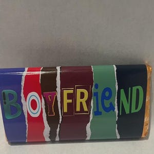 Op de afbeelding: Een chocoladereepverpakking met het woord "BOYFRIEND" in kleurrijke letters. De verpakking heeft een blauwe, rode, bruine, bordeauxrode, groene en marineblauwe achtergrond met een gescheurd papiereffect.