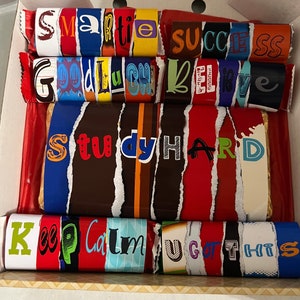 May include: A box of chocolate bars with motivational messages. The bars are arranged in a stack and have different colours and designs. The messages on the bars include "Smartie Success", "Good Luck", "Believe", "Study Hard", "Keep Calm", and "You Got This".