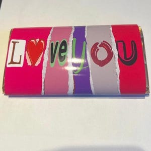 Op de afbeelding: Een verpakte chocoladereep met de woorden "LOVE YOU" in een kleurrijk ontwerp. De letters zijn in verschillende kleuren, waaronder roze, rood, groen en paars, met een hart dat de "O" in "LOVE" vervangt.