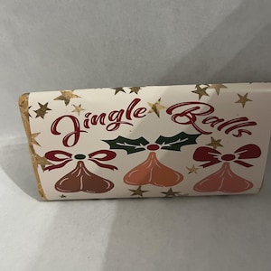 May include: A rectangular package with the words "Jingle Balls" in red script, decorated with gold stars, holly, and three bell-shaped ornaments in shades of brown and pink. The package has a festive, holiday theme.