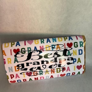 Op de afbeelding: Een witte chocolade reep met een kleurrijk patroon van de woorden "grandpa" en harten. De tekst "Best grandpa ever" is in zwart op de reep gedrukt.