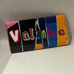 Op de afbeelding: Een rechthoekige chocoladereep met het woord "Valentine" op de voorkant. De reep is verdeeld in kleurrijke secties van roze, zwart, blauwgroen, blauw en oranje. De letter "V" is wit met een rood hart.