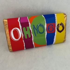 Op de afbeelding: Een verpakte chocoladereep met een kleurrijk ontwerp. De verpakking toont de woorden "OH NO 6" in wit en blauw, met een rode, roze, oranje, groene, blauwe en gele achtergrond. De uiteinden van de reep zijn verpakt in goudfolie.