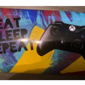 Puede incluir: Un objeto rectangular con el texto "EAT SLEEP REPEAT" en letras negras. Un mando de Xbox negro está a la derecha. El fondo tiene un diseño abstracto colorido con azul, rojo y amarillo.